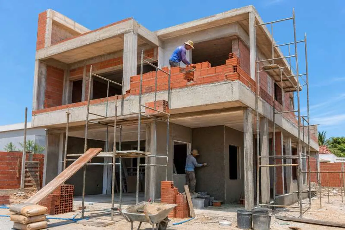 Casa em construção em Saco Grande, com estrutura de tijolos, andaime e operários trabalhando na obra